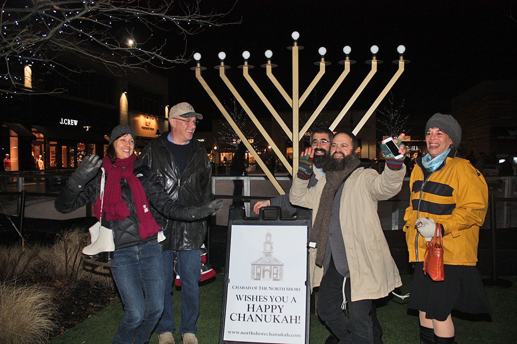 MEMBERS of Chabad of the North Shore were excited to help send the message of light during a Chanukah celebration for the community held at MarketStreet Lynnfield last Thursday on the third night of Chanukah. From left: Sheryl Perlow, Neil Donnenfeld, Rabbi Nemechia Schusterman, Rabbi Yossi Lipsker and Mara B. Cohen dance near the nine-foot tall menorah situated near the ice skating rink on the green.  (Maureen Doherty Photo)